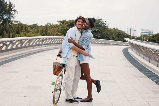 Plano completo de la joven pareja étnica abrazando a la bicicleta en el puente