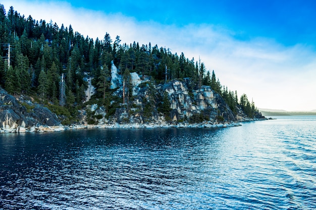 Plano general de un mar azul claro cerca de una montaña cubierta de pinos bajo un cielo despejado