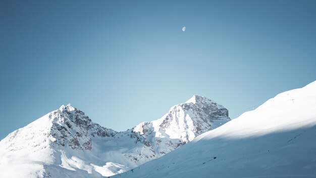 Plano general de montañas cubiertas de nieve bajo un cielo azul claro con media luna