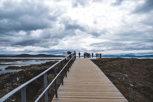 Plano general de un paseo marítimo con barandilla y turistas, con vistas a un lago en un día nublado