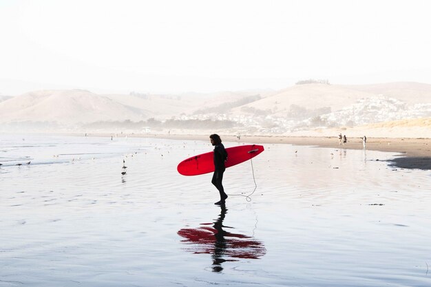 Plano general de un surfista masculino con traje de surf sosteniendo una tabla de surf en la orilla del mar