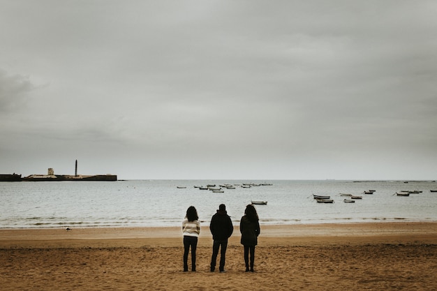 Plano general de tres personas de pie cerca de la orilla del mar con pequeñas embarcaciones flotando en el mar