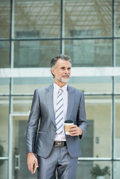 Plano medio del ejecutivo de negocios inteligente en ropa formal de pie fuera del edificio de oficinas para tomar su café para llevar