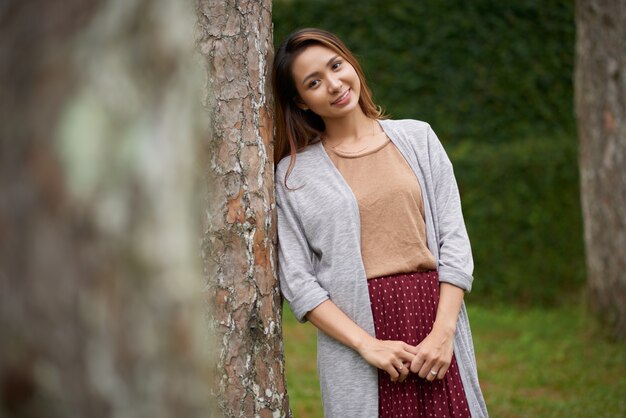 Plano medio de la joven mujer asiática apoyándose en el árbol y posando para una foto en el parque