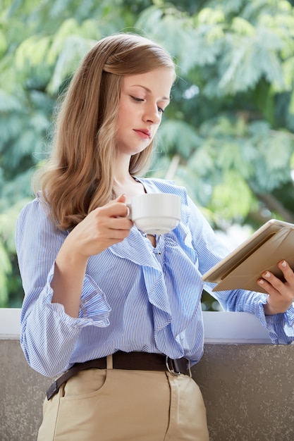 Plano medio de la joven rubia de pie en el balcón con una taza de café y una tableta de lectura