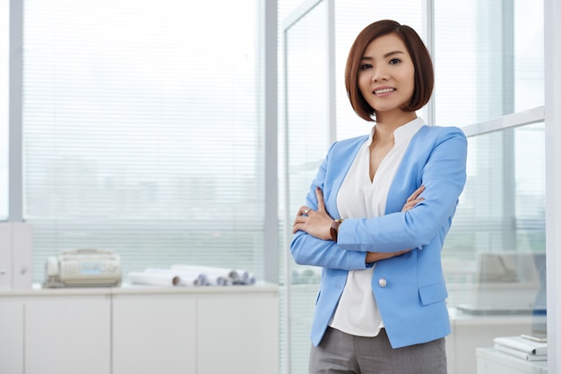 Plano medio de la mujer de negocios asiática de pie en la oficina con los brazos cruzados