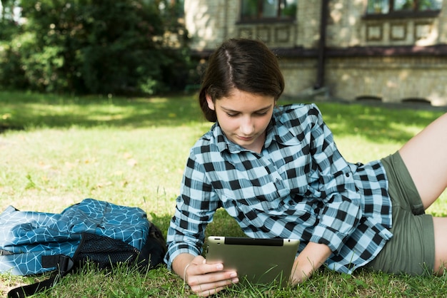 Plano medio de niña de la escuela usando tableta sentado en el césped