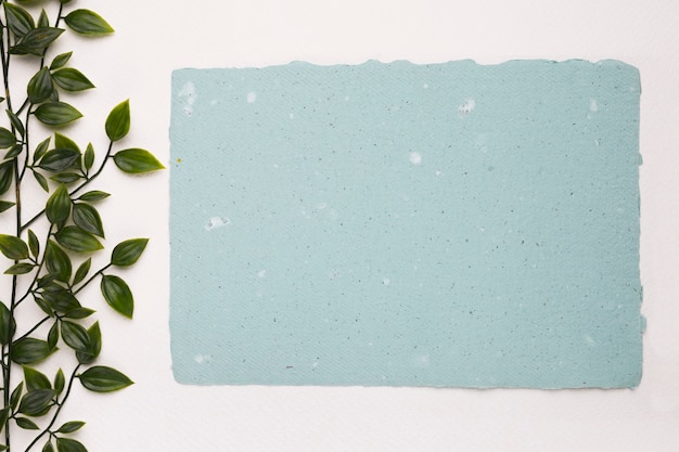 Una planta verde artificial cerca del papel de textura azul en blanco sobre fondo blanco.