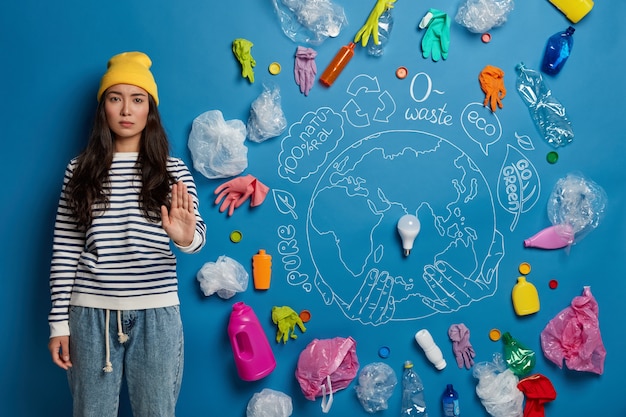 Foto gratuita sin plástico y contaminación ambiental. mujer asiática seria tira de la palma hacia adelante, usa sombrero amarillo, jersey a rayas y pantalones de mezclilla, pide no contaminar nuestro planeta