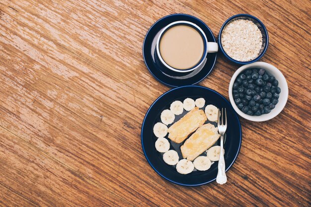 Plátano; pan tostado; bayas azules Taza de muesli y café sobre fondo con textura de madera