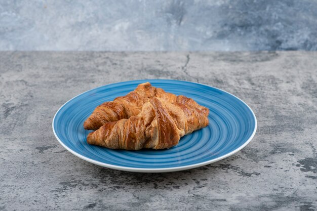 Un plato azul de dos croissants recién hechos en una mesa de piedra.