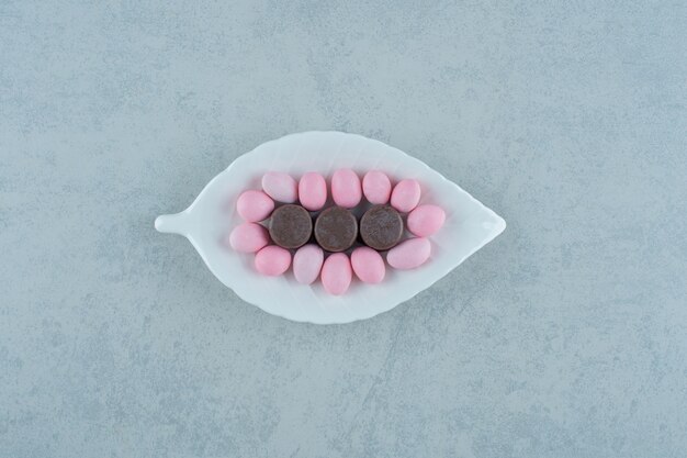 Un plato blanco con dulces caramelos de color rosa y galletas de chocolate en la superficie blanca
