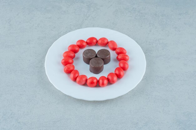 Un plato blanco con dulces caramelos rojos y galletas de chocolate sobre superficie blanca