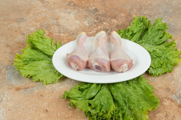 Un plato blanco con muslo de pollo crudo y lechuga sobre superficie de mármol.