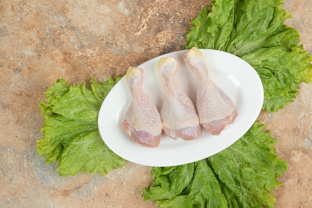 Un plato blanco con muslo de pollo crudo y lechuga sobre superficie de mármol