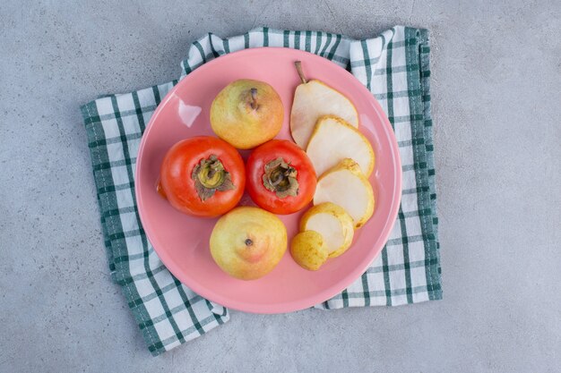 Un plato de frutas con peras y caquis sobre una toalla, sobre fondo de mármol.