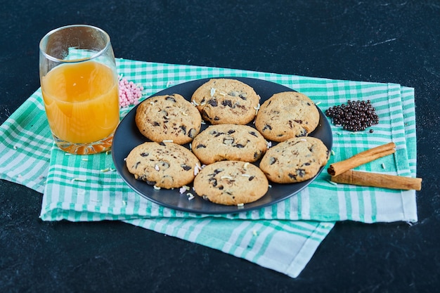 Un plato de galletas de chispas de chocolate y un vaso de jugo de naranja en la mesa oscura con canela