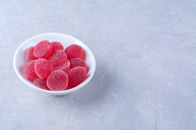 Un plato hondo blanco lleno de caramelos de gelatina de frutas azucaradas rojas sobre fondo gris. Foto de alta calidad