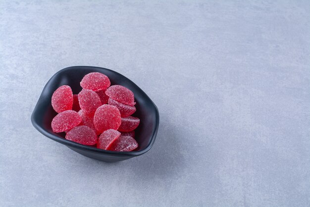 Un plato hondo negro lleno de caramelos de gelatina de frutas azucaradas rojas en la mesa gris.