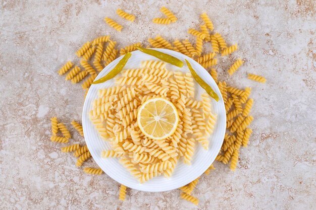 Plato de macarrones rodeado por un montón de pasta cruda y cubierto con una rodaja de limón sobre una superficie de mármol.