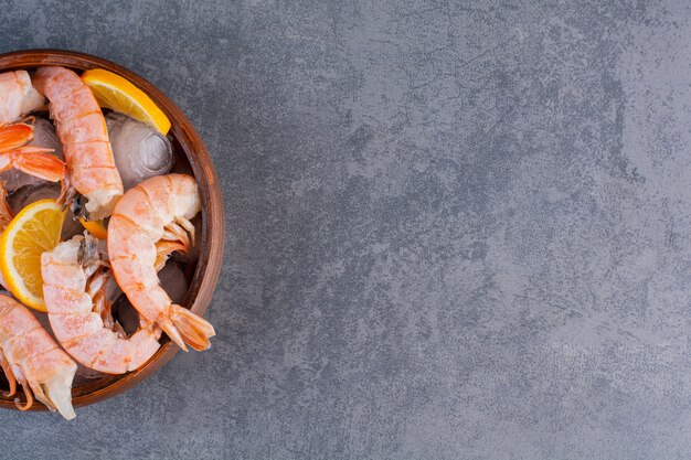 Un plato de madera de deliciosos camarones con cubitos de hielo y rodajas de limón sobre un fondo de piedra.