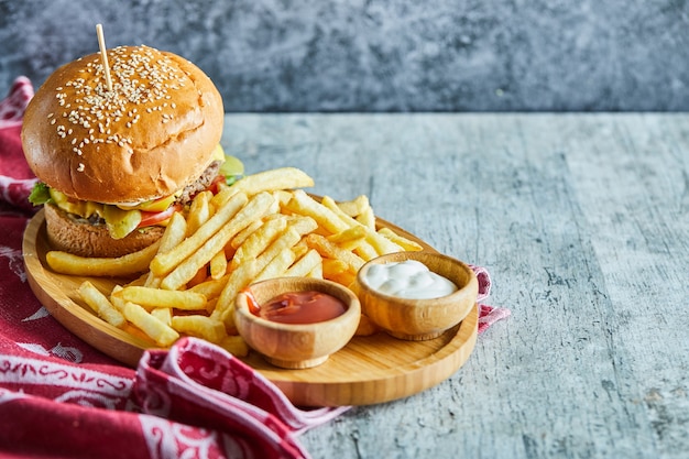 Un plato de madera lleno de hamburguesa, patatas fritas con salsa de tomate y mayonesa sobre la mesa de mármol.