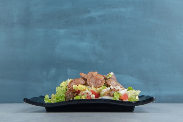 Un plato negro de pollo frito, carne y ensalada de verduras.