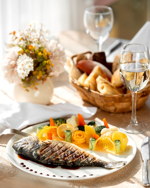 Un plato de pescado a la parrilla servido con verduras hervidas y rodajas de naranja.