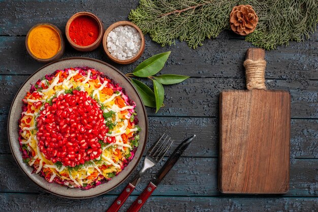 Plato de plato de Navidad de vista superior con granada, zanahorias, patatas junto a la tabla de cortar de madera, tazones de fuente de diferentes especias ramas de abeto con conos sobre la mesa