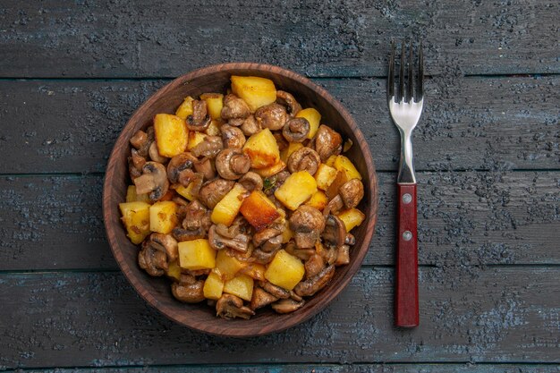 Plato de visión cercana superior y cuenco de tenedor con apetitosas papas y champiñones junto al tenedor en la mesa oscura