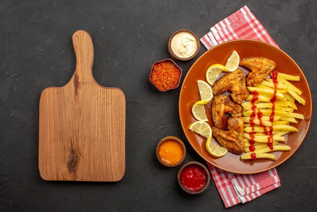 Plato de vista superior de alitas de pollo de comida rápida, papas fritas con limón y salsa de tomate y tazones de salsas y especias sobre un mantel a cuadros rosa-blanco junto a la tabla de cortar de madera