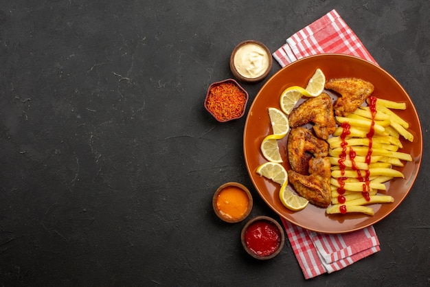 Plato de vista superior de alitas de pollo de comida rápida, papas fritas con limón y salsa de tomate y tazones de salsas y especias sobre un mantel a cuadros rosa-blanco en el lado derecho de la mesa negra