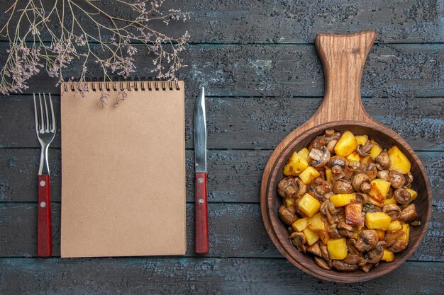 Plato de vista superior y cuaderno un plato de patatas y champiñones en la tabla de cortar y un cuaderno entre tenedor y cuchillo