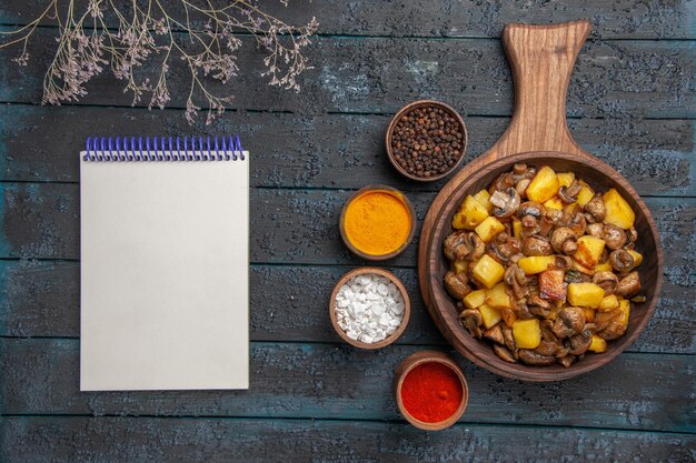 Plato de vista superior y especias un plato de papas y champiñones en la tabla de cortar y especias coloridas a su alrededor junto al cuaderno y ramas