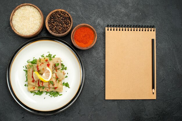 Plato de vista superior y especias repollo relleno con salsa de limón y hierbas y tazones de especias de colores, arroz y pimienta negra en la mesa junto al cuaderno de crema y lápiz