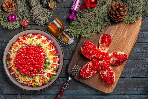 Plato de vista superior y plato de especias con granada junto a la botella de tenedor de granada con aceite en la tabla de cortar y ramas de árbol con conos y juguetes de árbol de Navidad