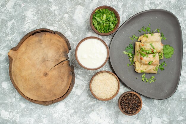 Plato de vista superior con plato de hierbas de repollo relleno junto a la tabla de cortar de madera y tazones de hierbas arroz crema agria y pimienta negra sobre la mesa