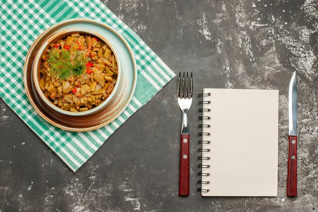 Plato de vista superior sobre el mantel blanco cuaderno tenedor cuchillo y plato de las apetitosas judías verdes con tomates en el tablero sobre el mantel a cuadros en la mesa oscura