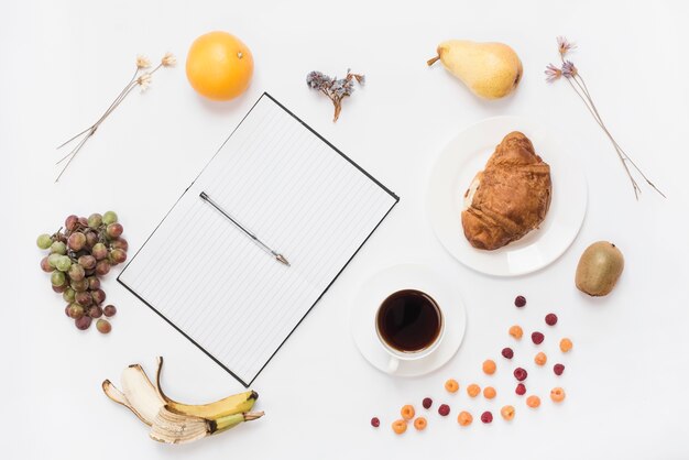 Pluma en un cuaderno abierto con taza de café; Croissant y muchas frutas sobre fondo blanco