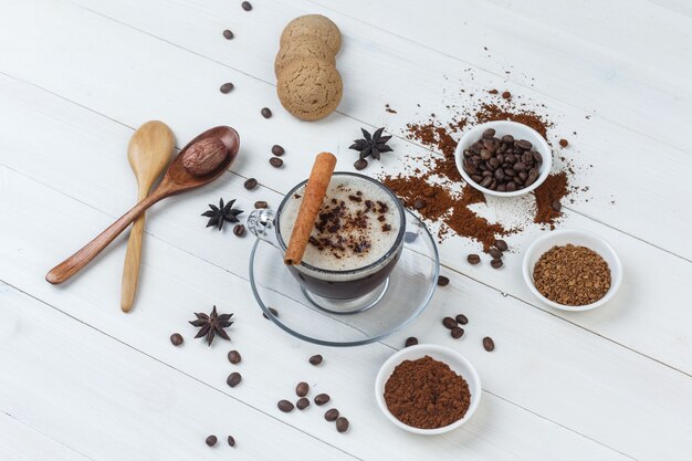 Un poco de café con granos de café, café molido, especias, galletas, cucharas de madera en una taza sobre fondo de madera, vista de ángulo alto.