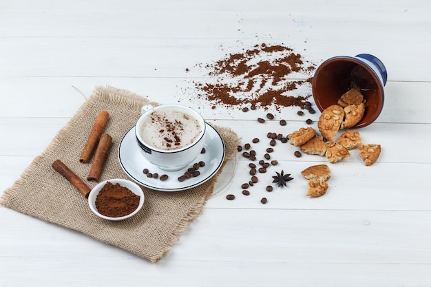 Un poco de café con granos de café, café molido, galletas, canela en una taza de madera y un pedazo de fondo de saco, vista de ángulo alto.