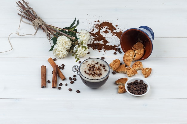 Un poco de café con granos de café, galletas, flores, canela en una taza sobre fondo de madera, vista de ángulo alto.