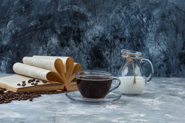 Un poco de café con granos de café, libro, leche en una taza sobre fondo de yeso y grunge, vista lateral.