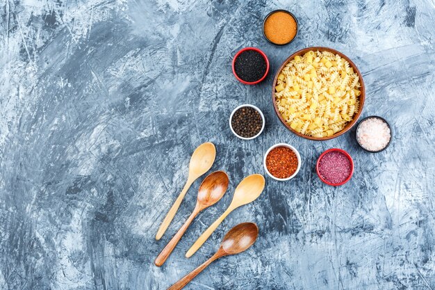 Un poco de pasta cruda con cucharas de madera, especias en tazones sobre fondo de yeso gris, vista superior.