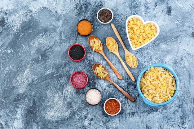 Un poco de pasta cruda con especias en tazones y cucharas de madera sobre fondo de yeso gris, vista superior.