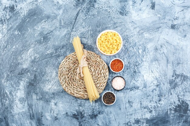 Un poco de pasta ditalini con espaguetis, especias en un recipiente sobre fondo gris de yeso y mantel de mimbre, vista superior.