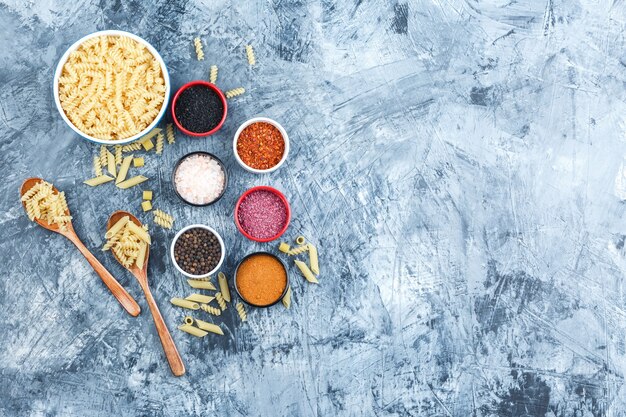 Un poco de pasta fusilli con especias en un tazón y cucharas de madera sobre fondo de yeso gris, vista superior.