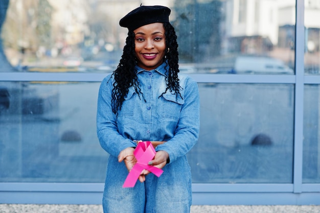 El poder de luchar contra las mujeres afroamericanas de moda con estilo en jeans y boina negra contra el edificio moderno con cinta rosa para el cáncer de mama