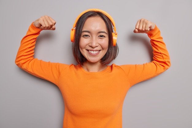 Poderosa mujer asiática segura de sí misma levanta los brazos, muestra los bíceps después del entrenamiento, sonríe, escucha agradablemente la pista de audio en los auriculares, usa un jersey naranja de manga larga, es fuerte, saludable, disfruta de la lista de reproducción de música