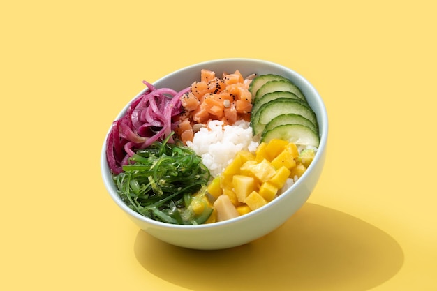 Poke bowl con arroz, salmón, pepino, mango, cebolla, ensalada de wakame, semillas de amapola y semillas de girasol aisladas sobre fondo amarillo.